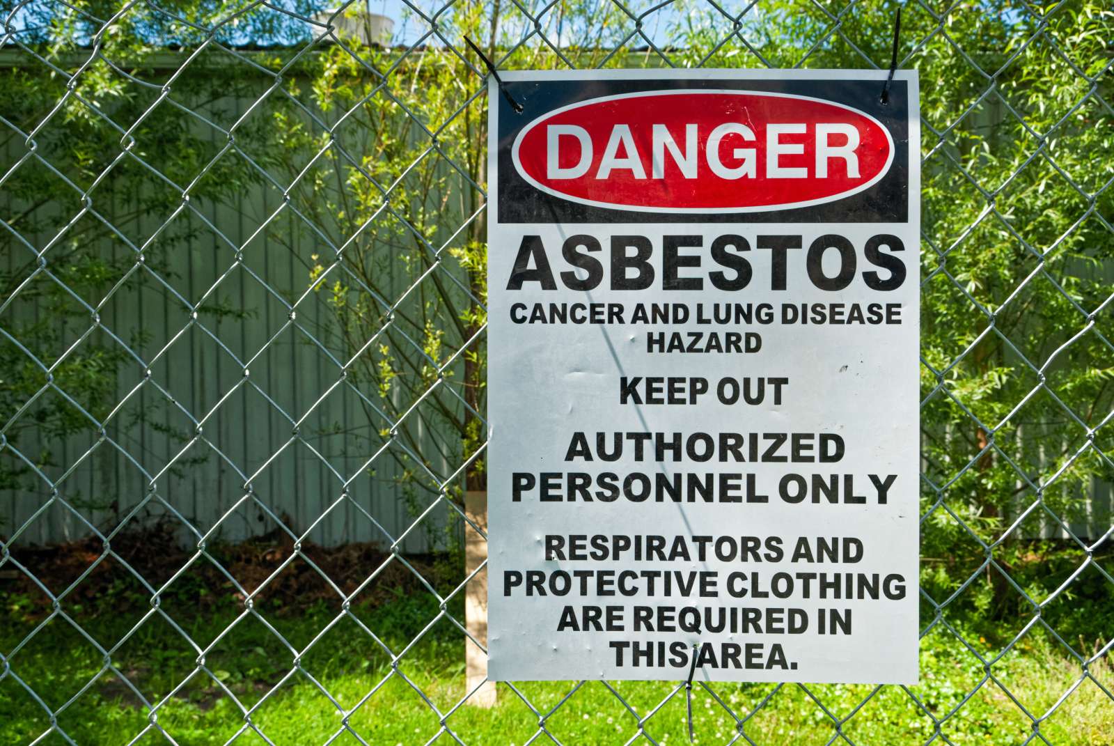 A sign is posted on a chain-link fence that says, "Danger: Asbestos. Cancer and lung disease hazard. Keep out. Authorized personnel only. Respirators and protective clothing are required in this area."