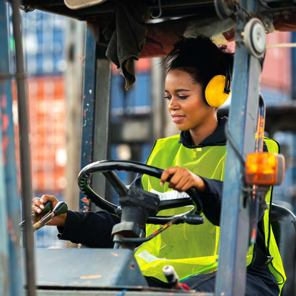 Person driving forklift in a warehouse while wearing hearing protection and a high-visibility vest