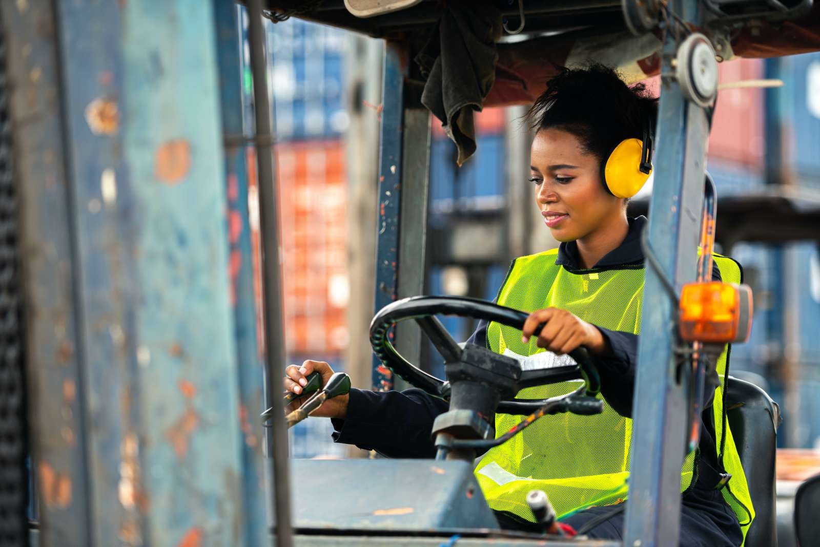Woman wears safety vest and hearing protection while operating a forklift