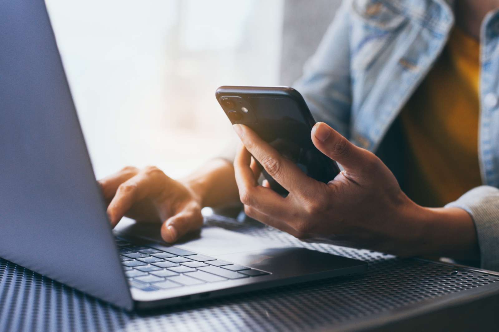 A person holds a smart phone in one hand while typing on a laptop with the other